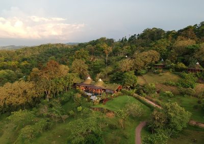 An aerial view of a lush, green landscape with dense trees and vegetation. In the midst of the forested area, there's a cluster of buildings with thatched roofs. One large structure appears to have an outdoor seating area, possibly a restaurant or gathering space. The scene is lit by the soft glow of the setting sun, casting a warm light over the landscape. A winding path snakes through the greenery, connecting different parts of the property rio celeste Costa Rica hotels