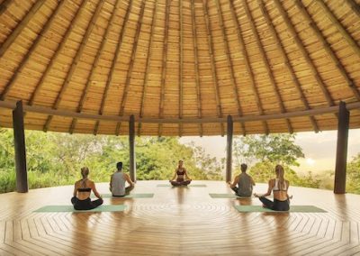A serene open-air pavilion with a thatched roof, set amidst nature. A group of six individuals sits in a circle on a patterned floor, engaged in meditation or yoga. They are evenly spaced, facing a central instructor who guides them. The setting sun outside casts a golden hue, enhancing the tranquil ambiance of the scene.