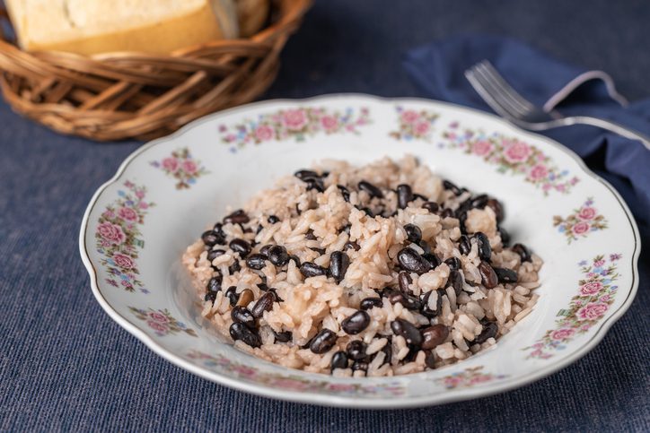 A plate with a floral design contains a Costa Rica food Gallo pinto serving of rice mixed with black beans, presenting a classic dish. In the background, there's a wicker basket holding slices of bread, all set against a textured blue tablecloth. A silver fork rests nearby, hinting at an upcoming meal.