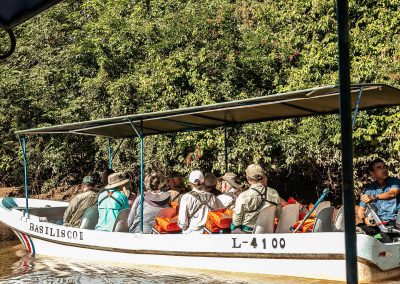 Passengers in safari attire riding a boat with a guide on a river, surrounded by lush greenery.