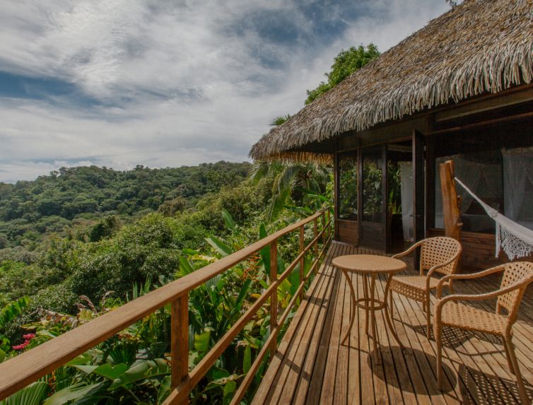 Wooden cabin with a thatched roof and a deck featuring chairs and a hammock, overlooking a lush green forest under a cloudy sky, perfect for where to stay in Costa Rica.