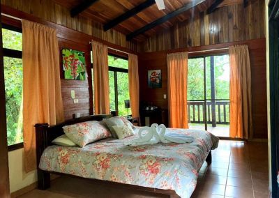 A cozy bedroom with a floral bedspread, wooden walls and flooring, and open curtains leading to a balcony surrounded by greenery.