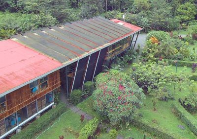 Aerial view of a large building with a rusty metal roof surrounded by lush green gardens in a tropical setting.