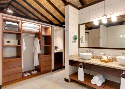 Luxurious bathroom with dual vessel sinks, mirrored wall, wooden cabinets, and an open wardrobe stocked with towels and a white bathrobe.