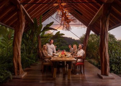 A family enjoying dinner in an open wooden pavilion surrounded by lush greenery at dusk.