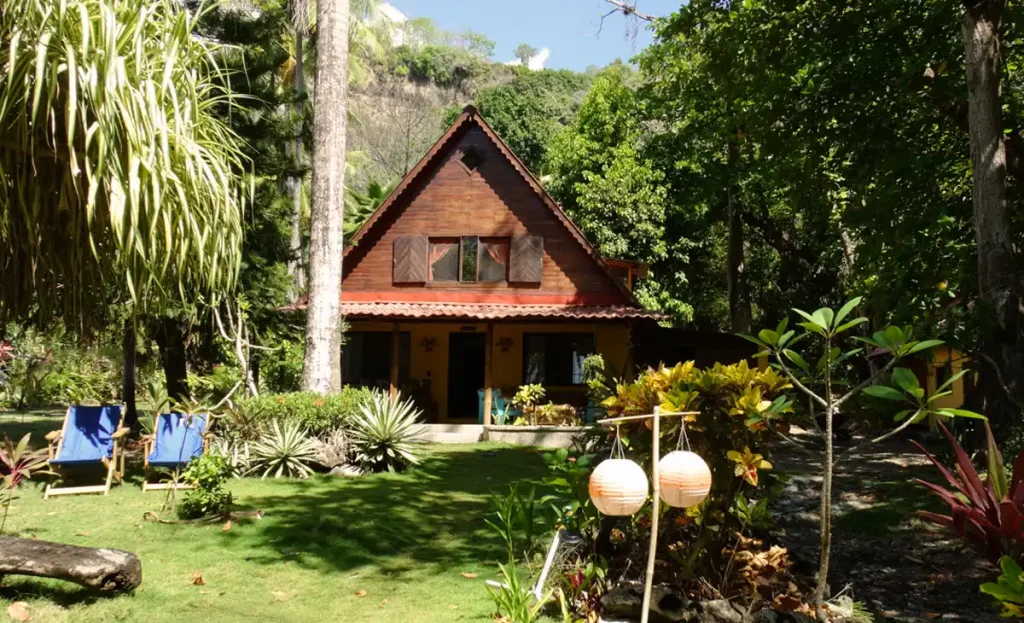 A quaint triangular-roofed cabin surrounded by lush greenery and tropical plants in Costa Rica, with two blue chairs on the grass and decorative lanterns hanging outside.