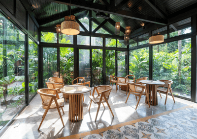 Interior of a glass-walled sunroom with lush greenery outside, featuring rattan chairs, round tables, and pendant lights.