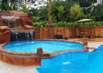 Tropical resort style swimming pool with a blue water, artificial waterfall and naturalistic rock features surrounded by lush greenery.