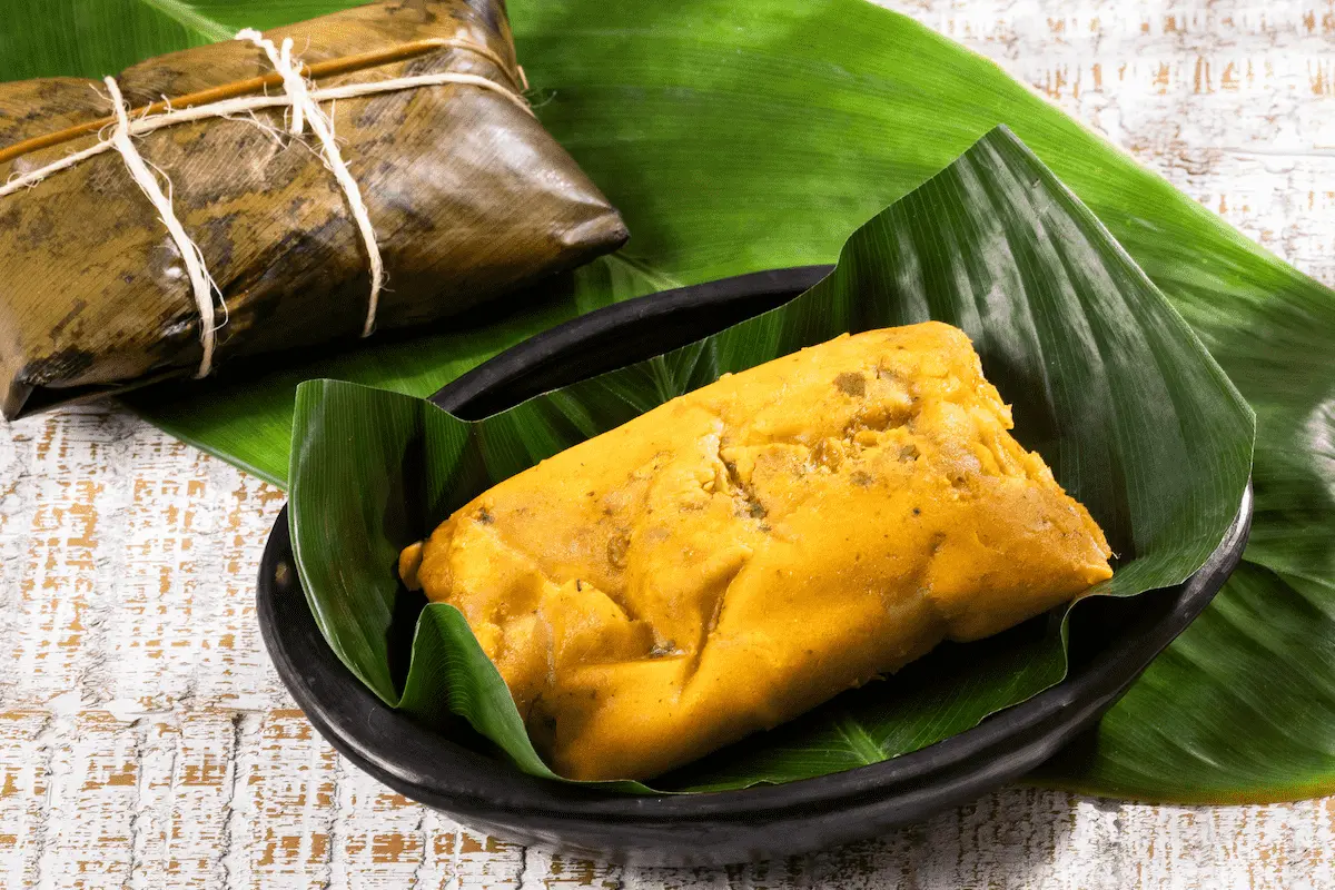 A traditional tamale is served on a green leaf in a black bowl, with another tamale wrapped in a leaf and tied with string placed behind it, also on a green leaf.