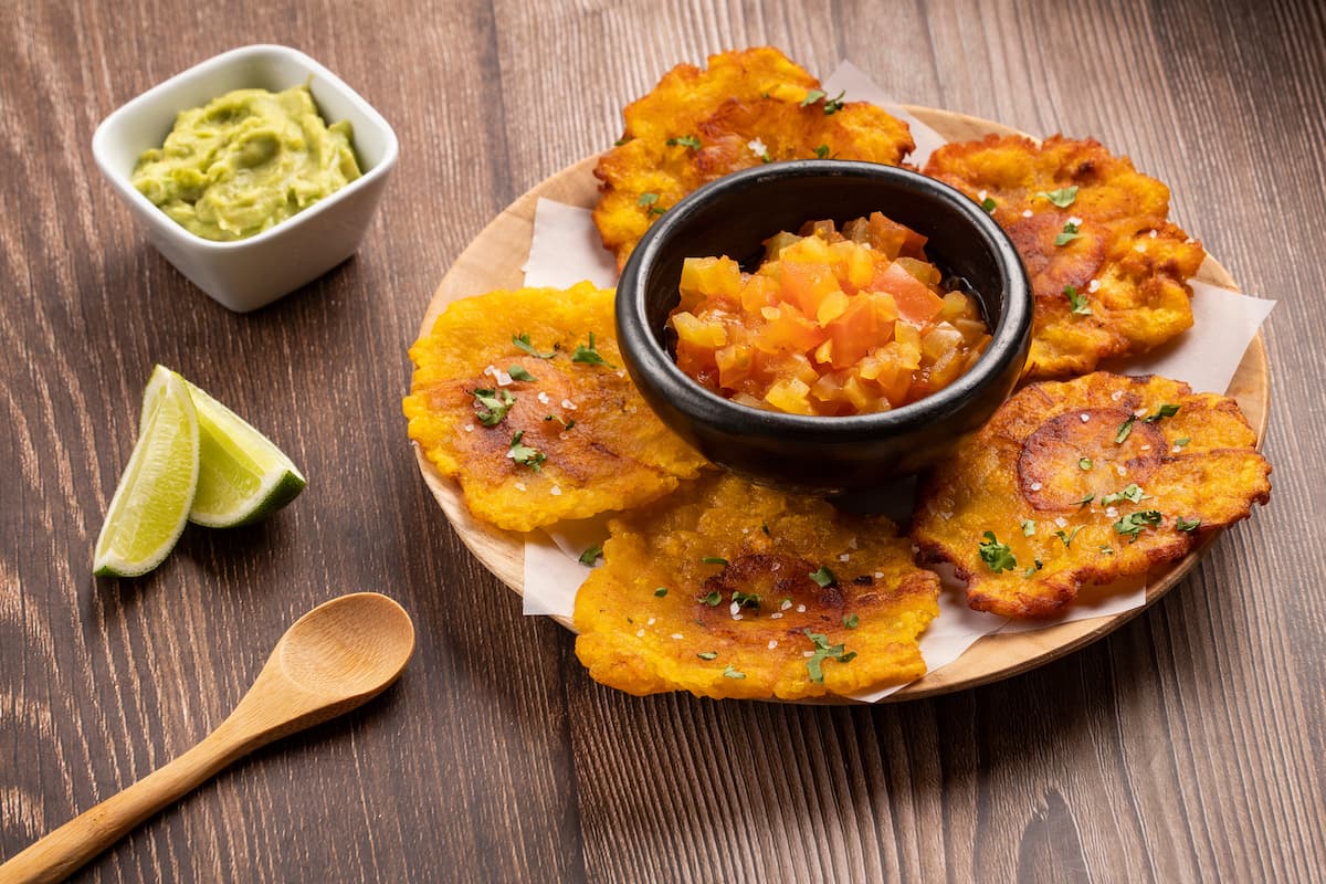 A plate of patacones with a bowl of tomato salsa in the center. A side of guacamole and lime wedges are nearby, along with a wooden spoon, on a wooden table.