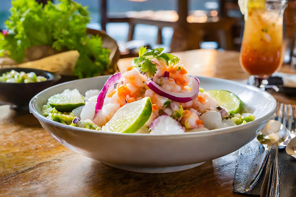 A bowl of ceviche Costa Rican food garnished with lime wedges and red onions sits on a wooden table, with a drink and salad in the background.
